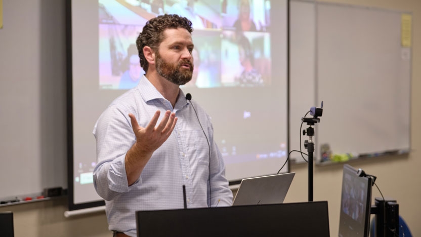 Lecturer at a podium with a computer and screen behind.