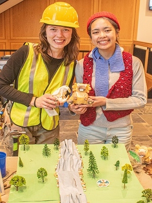 Two people stand in front of a model environment with trees and a river, smiling at the camera and holding objects.