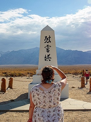 A person stands before a monument written in Japanese, looking towards the monument and away from the camera. In the background you can see mountains and a blue sky with white clouds.