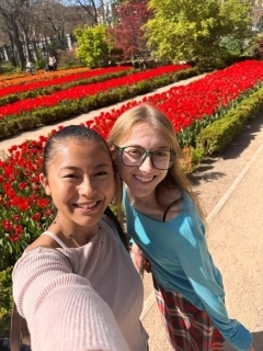 Two students smiling in a selfie in front of red flowers beds