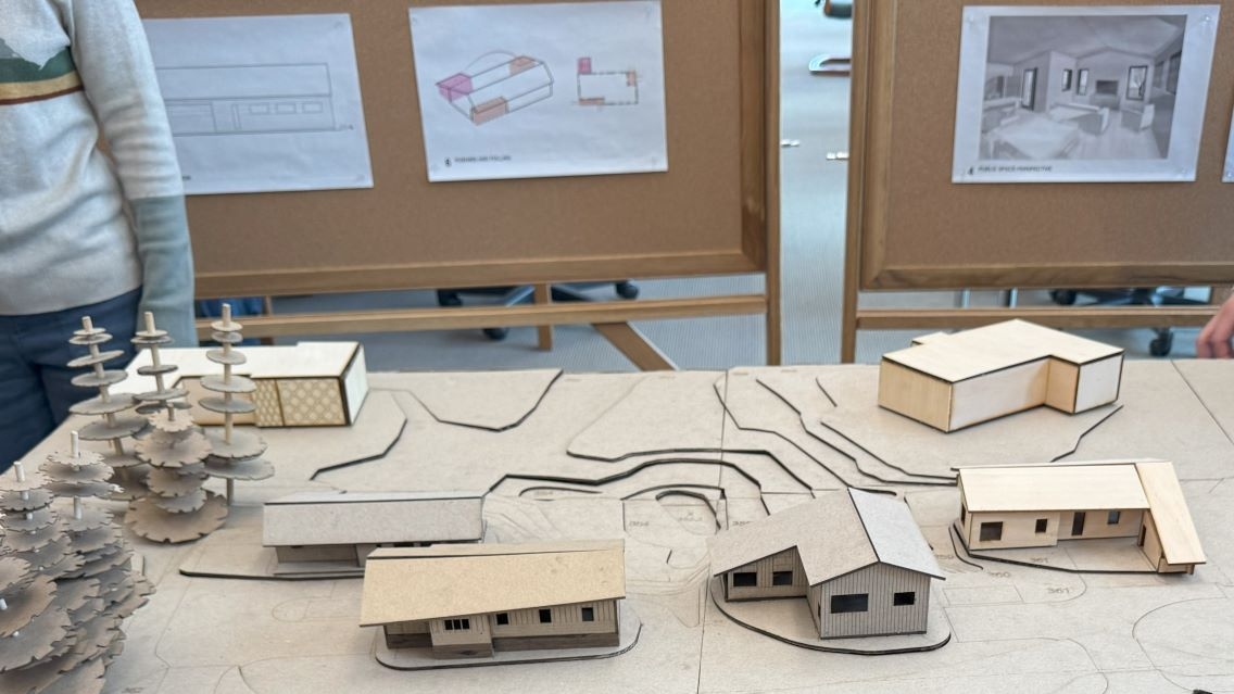 Table-top architectural model of several residential structures with architectural drawing and rendering on bulletin boards in the background.