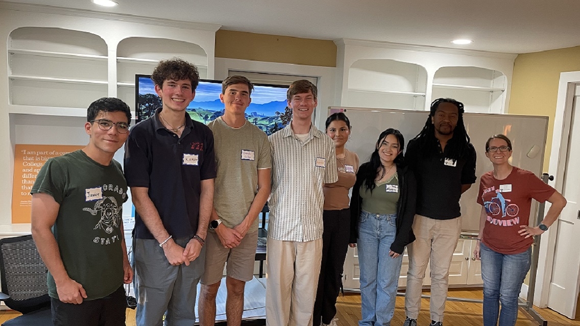 8 people stand indoors in a line and smile at the camera. They are standing in front of white shelves, a large computer screen, and a whiteboard.