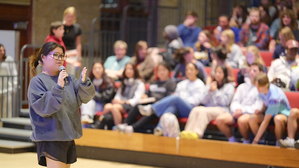 A person wearing glasses, a grey hoodie, and black shorts stands and speaks in to a microphone in front of a large crowd sitting in stands.