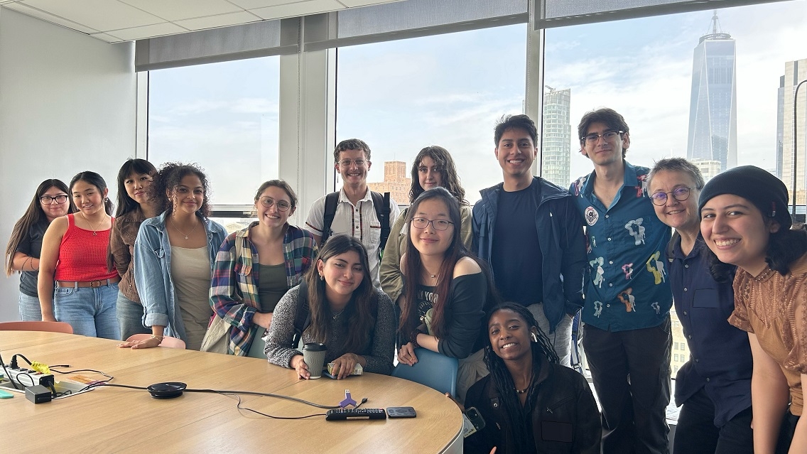 14 people stand indoors in a line and smile at the camera. They are standing in front of a table, and skyscrapers can be seen out a window behind the group.