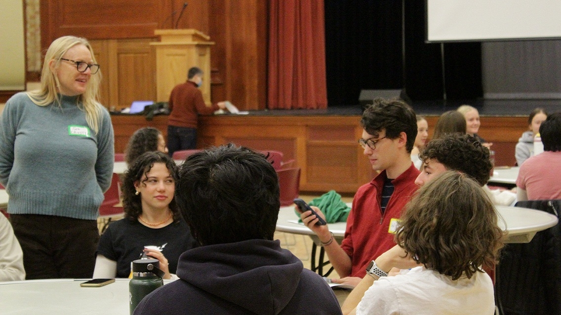 5 people sit at a table indoors and look at a person holding a phone and speaking. Another person stands near the table listening. In the background you can see a stage and podium.