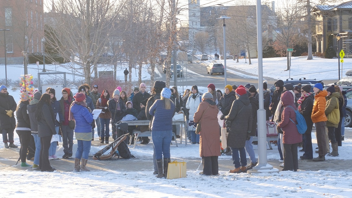 A large group of people stand outside in the snow and on a sidewalk. Musical instruments can be seen on the ground.
