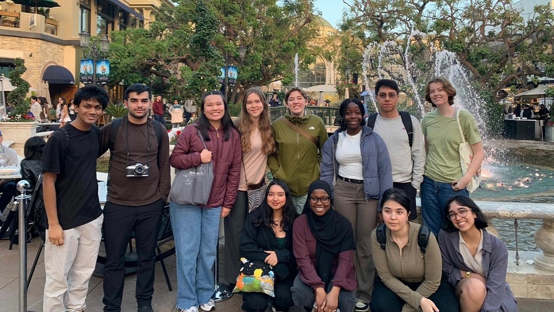 12 people stand outdoors in a line and smile at the camera, in front of a fountain and yellow building.