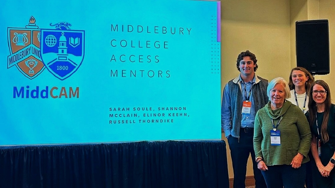 Four people smile at the camera while standing by a large computer screen that reads "Middlebury College Access Mentors".
