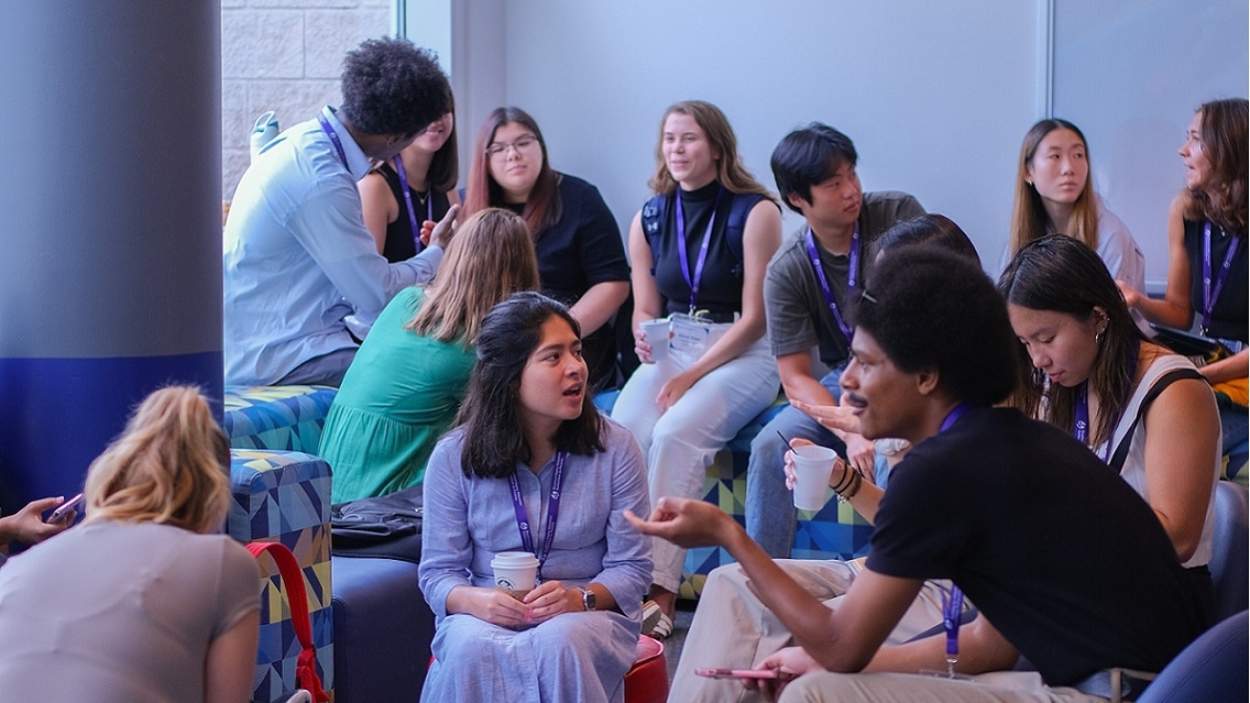 13 people sit on indoor furniture and speak in small groups, dressed nicely and wearing nametags around their necks. Some are holding drinks.