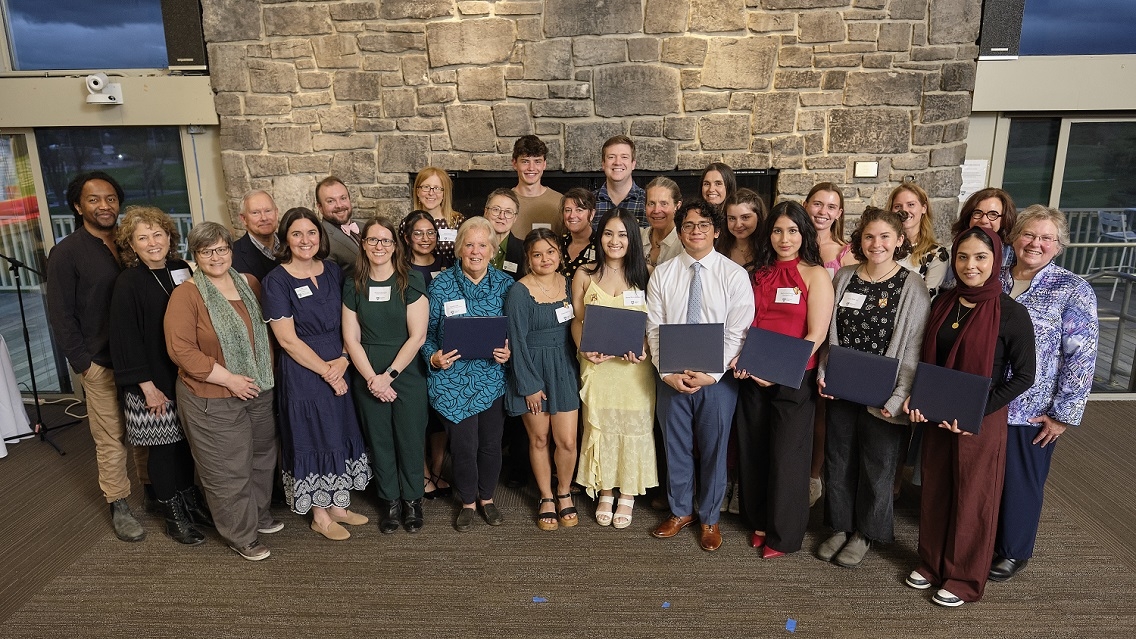 A large group of people stand indoors and smile at the camera. They are standing in front of a stone fireplace. Some are holding blue awards.
