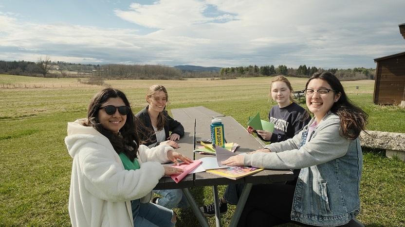 Four people sit at a table folding colored construction paper and smiling at the camera. A water bottle with the word "DREAM" can be seen on the table.