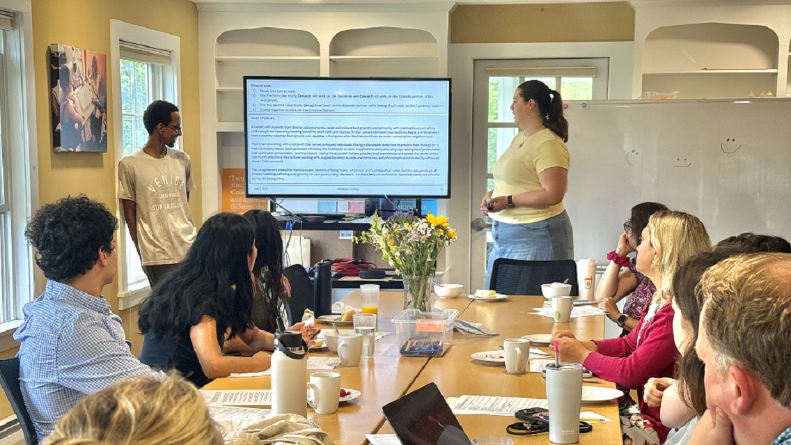 A group of students and faculty sit around a table looking up at a presentation on a large video monitor. Two presenters stand at the front of the table, also looking at the presentation on the video monitor.