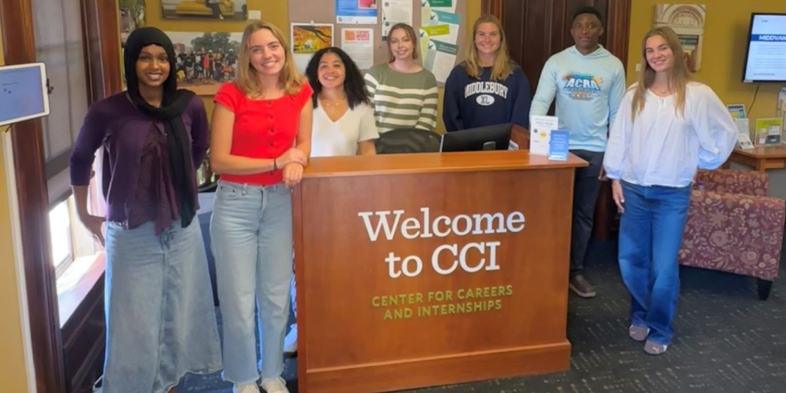 Group of seven students posing beside and behind the reception desk at CCI. A sign on the desk reads, "Welcome to CCI"