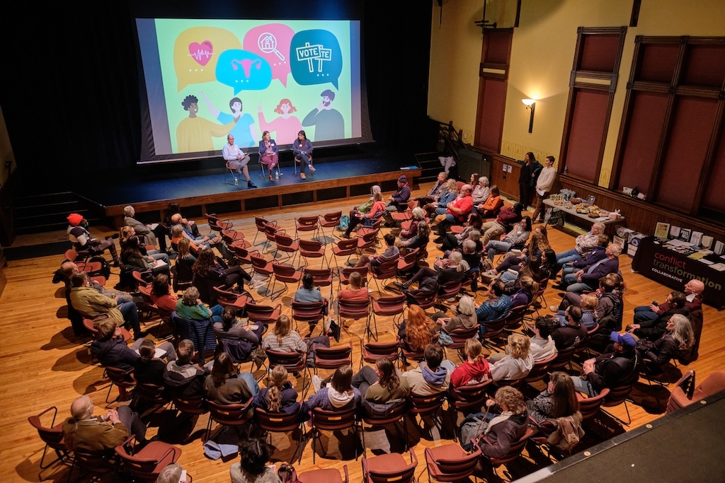 a photo taken above a large group of people sitting in concentric semi-circles, listening to a panel of presenters who sit in front of a large screen.