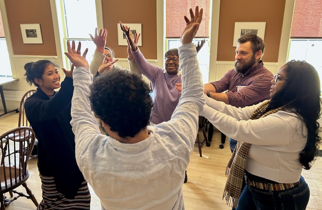 Five adults of different ethnicities stand in a circle looking at each other with their hands up
