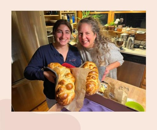 Two women stand together, facing the camera and holding giant loaves of freshley cooked bread. They are both smiling proudly.
