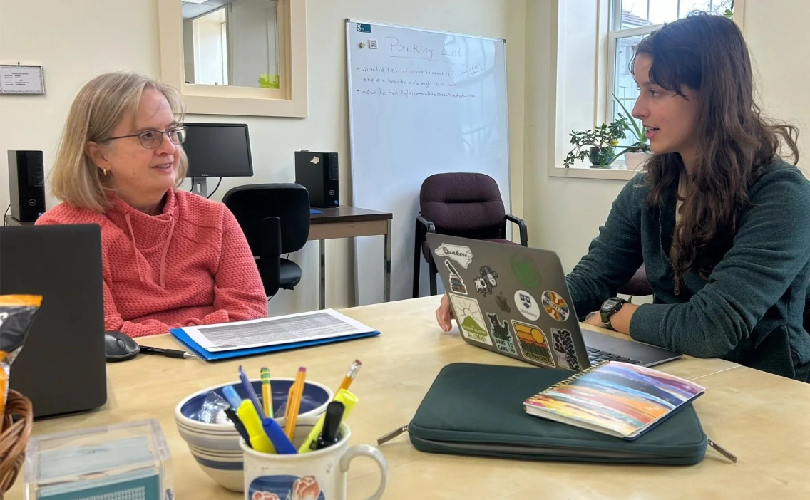 A young woman with long brown hair sits at a table in front of her laptop, and speaks to an older woman sitting opposite her. Both have warm expressions on their faces.