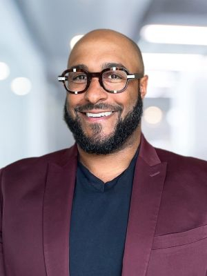 headshot of MAA Director nominee Andrew Plumley wearing glasses, blue shirt, and a red suit jacket