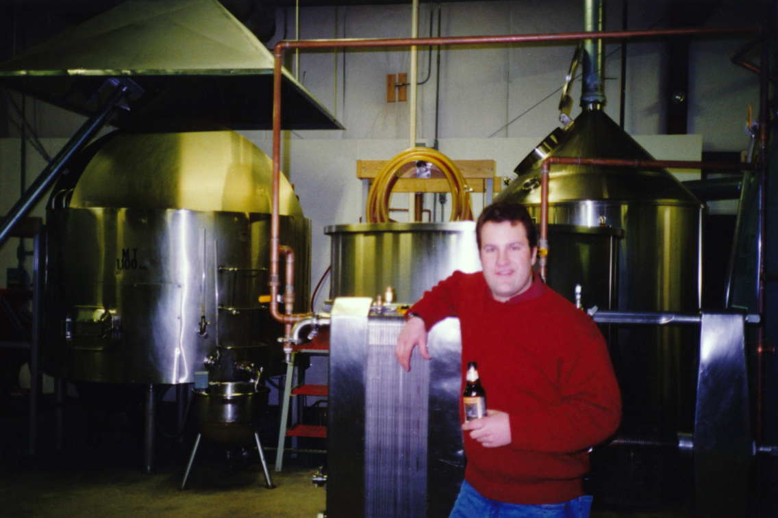 Rob Tod holds an Allagash White and poses with the original Allagash Brewing brewery system.