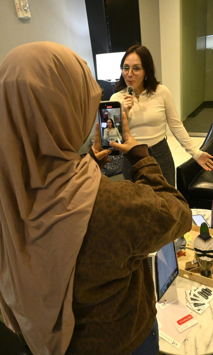 A person in a brown shirt and brown head scarf videotapes a young woman holding a microphone. She is wearing an ivory top and have shoulder-length brown hair.