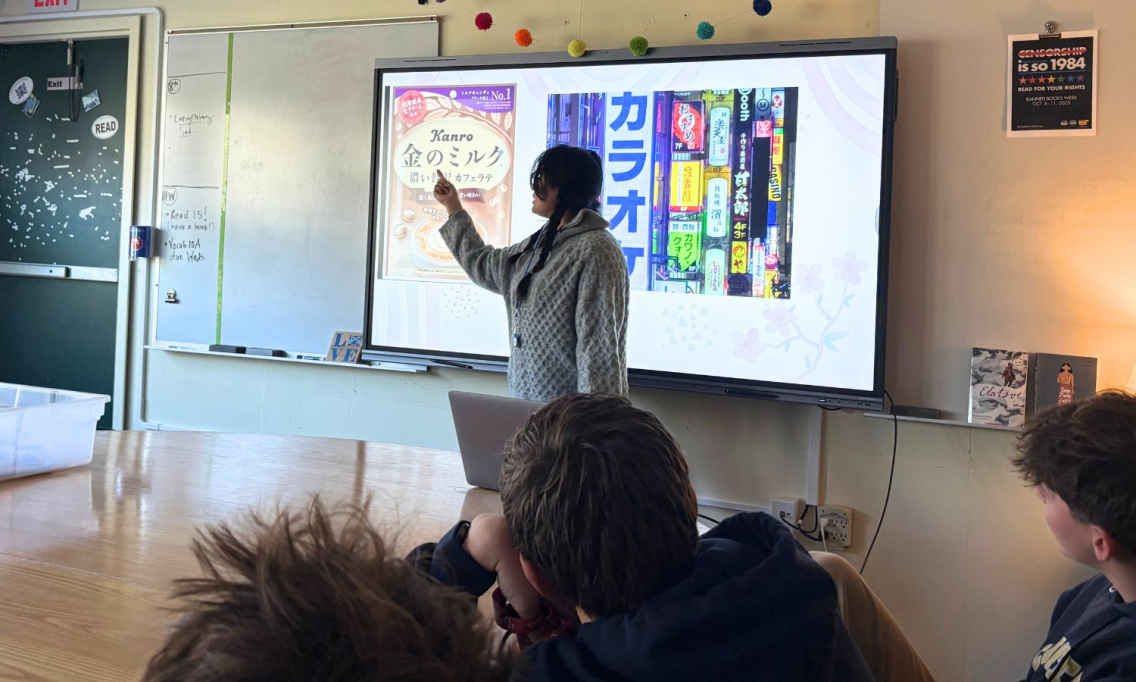 A young woman stands at the front of a classroom, pointing to the board behind her. You see the backs of student heads in the forefront of the photo.