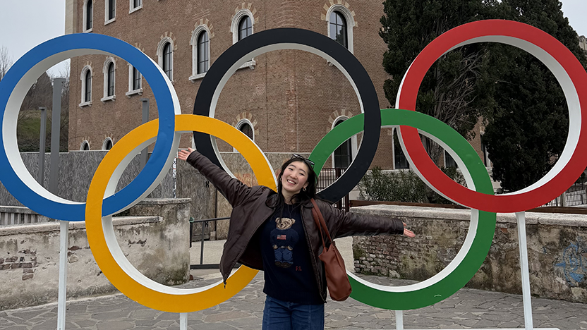 Ting Cui stands smiling before the Olympic Rings in Italy, arms raised over her head.