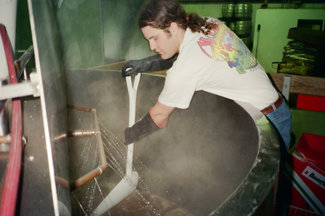 Rob Tod leans over a vat of hops and barley and uses a shovel to mix it up.
