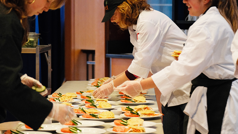 Students in Dolci kitchen uniforms plate chicken, tomato and cucumber on a large array of dishes