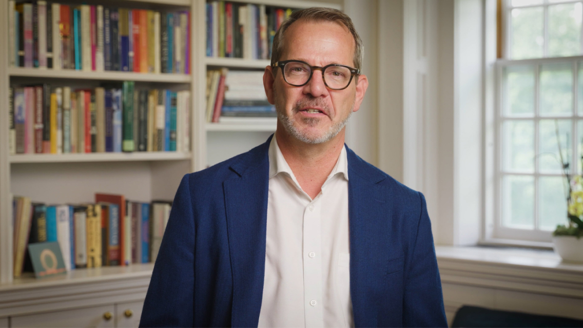 Still screen of Ian Baucom speaking in his office with a bookshelf in the background.