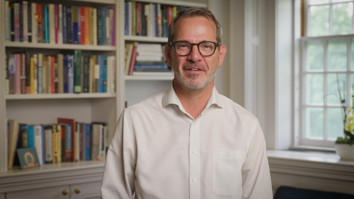 Middlebury president Ian Baucom wearing a white dress shirt speaks into a camera.