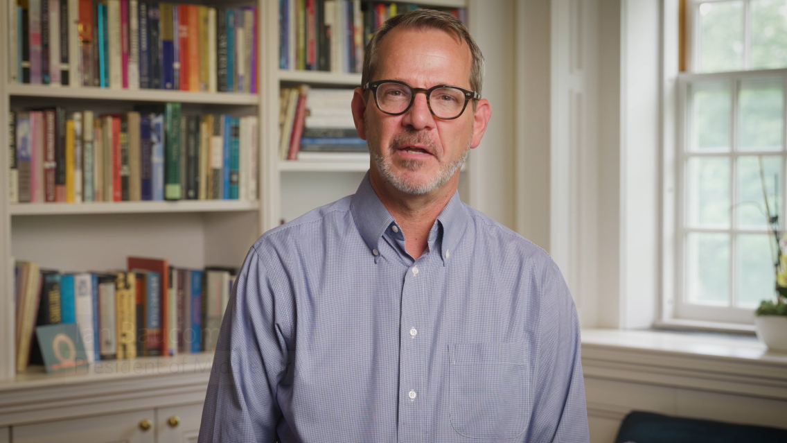 Middlebury president Ian Baucom wearing a blue dress shirt speaks into a camera.