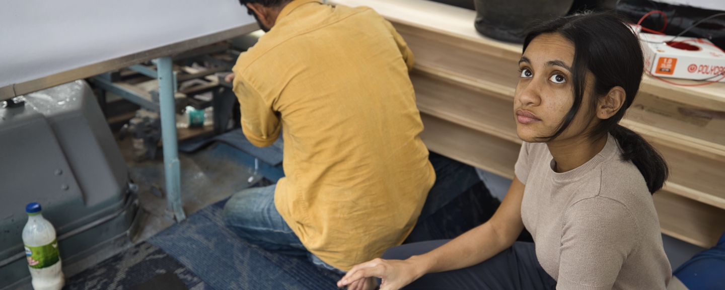 A woman wearing a beige shirt with her black hair pulled back in a ponytail looks up out of frame. Behind her, someone wearing a yellow shirt crouches down and works on an engine.