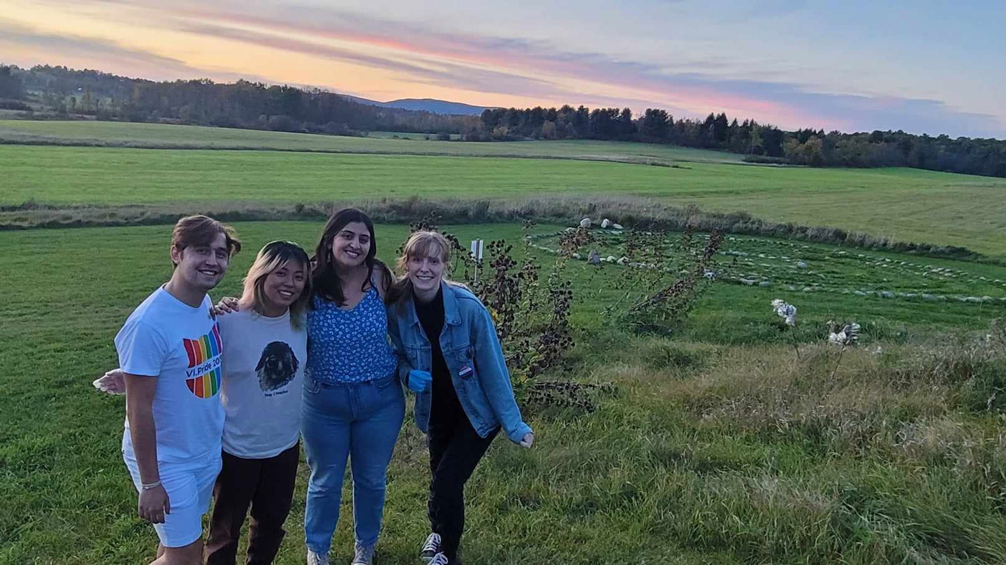 Four students stand arm in arm in a field at sunset on a spring evening.