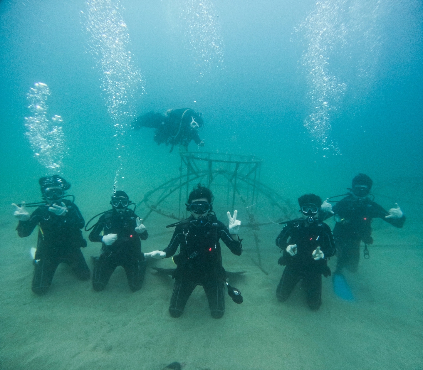 A group of scuba divers post underwater holding up peace signs as they work together on coral reef restoration project.