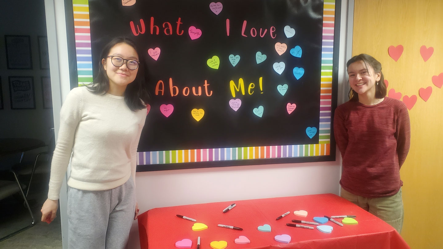 Two students stand in front of a poster tat reads "What I Love About Me!"