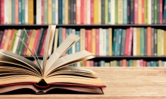 An open book with fanned pages sits on a wooden table in front of a packed shelf with multi-colored book spines