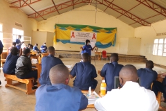 A woman in a blue shirt and mask stands in the center of a classroom addressing a group of circled up individuals in similar outfits.