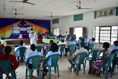 Standing at the front of a small gym with a stage, a teacher addresses a large group of students sitting in green lawn chairs.