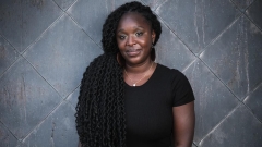 Masiray Kamara, wearing a black t-shirt and silver necklace, poses for a portrait in front of a gray, tiled, wall.