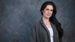 Leila Barghouty, wearing a white t-shirt and gray cardigan, poses for a portrait in front of a dark gray, tiled, wall.