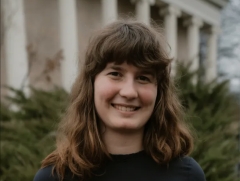 Wearing a navy sweater, Elisabeth Sundberg poses for a photo in front of a green bush and white, marble, columns.