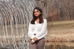 Ariha Shahed, wearing a white sweater and brown pants, poses in front of a small pond.