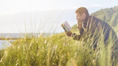 Picture of a man on a bench reading next to a body of water and tall green grass