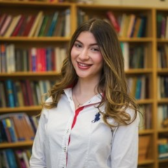 A woman stands smiling in a white shirt in front of a bookcase.