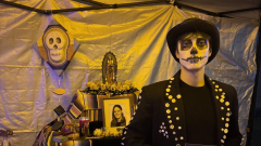 Ryan Ulen in traditional Dia de Muertos attire with face painted, stands in front of an ofrenda holding a framed photograph