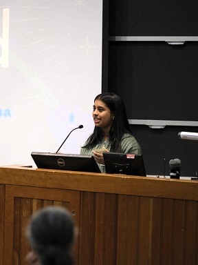 A person stands and speaks into a microphone, presenting research.