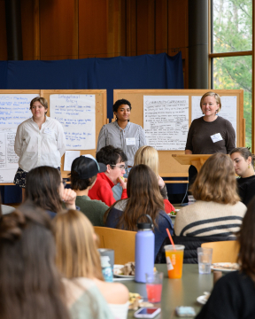 Student Ella and Staff members Reilly and Minna stand in front of brainstorming white boards set up in Atwater Dining Hall with a crowd in front of them