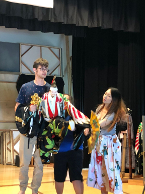 A tall young man participates in a cultural theater production. He holds a puppet.