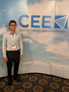 A young man in black dress pants and a grey button down stands smiling in front of a "CEEK Working Together. Advancing Efficiency" wall. 