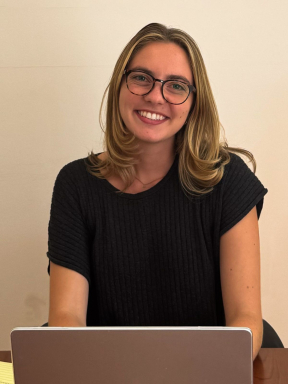A young woman with long light brown hair and a black short-sleeve shirt sits behind her laptop and smiles confidently at the camera.